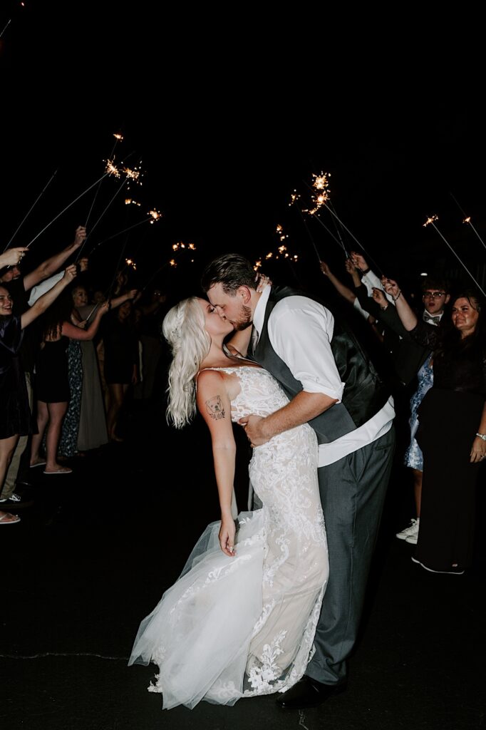 A bride and groom kiss one another while outside during their exit from their wedding ceremony, guests around them all hold sparklers and cheer