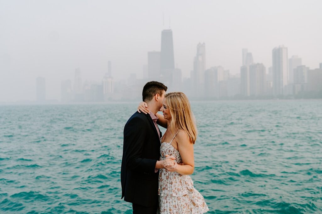A woman smiles while dancing with a man in front of Lake Michigan and a hazy Chicago skyline behind them during their engagement session