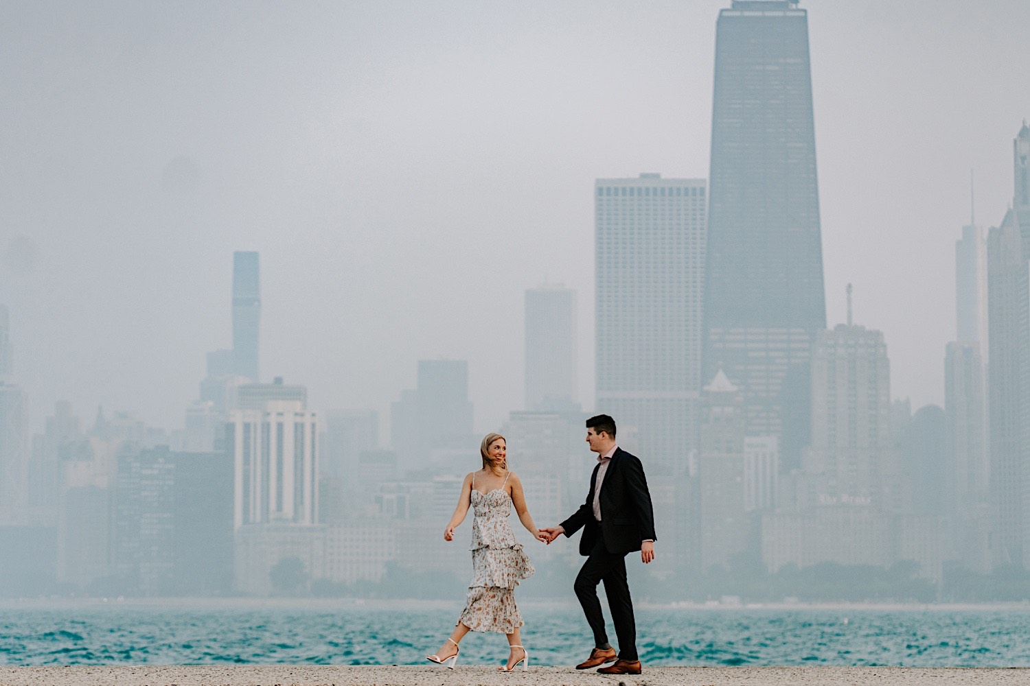 A man and woman walk hand in hand in front of Lake Michigan and the Chicago skyline behind them while the woman smiles back at the man