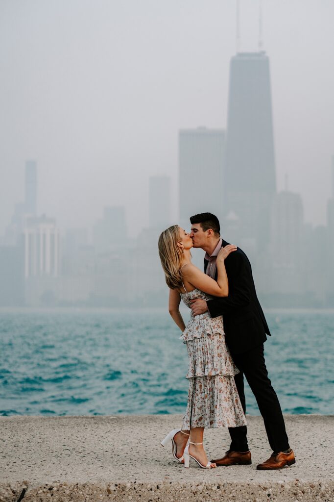 A man and woman kiss one another while standing in front of Lake Michigan and a hazy Chicago skyline