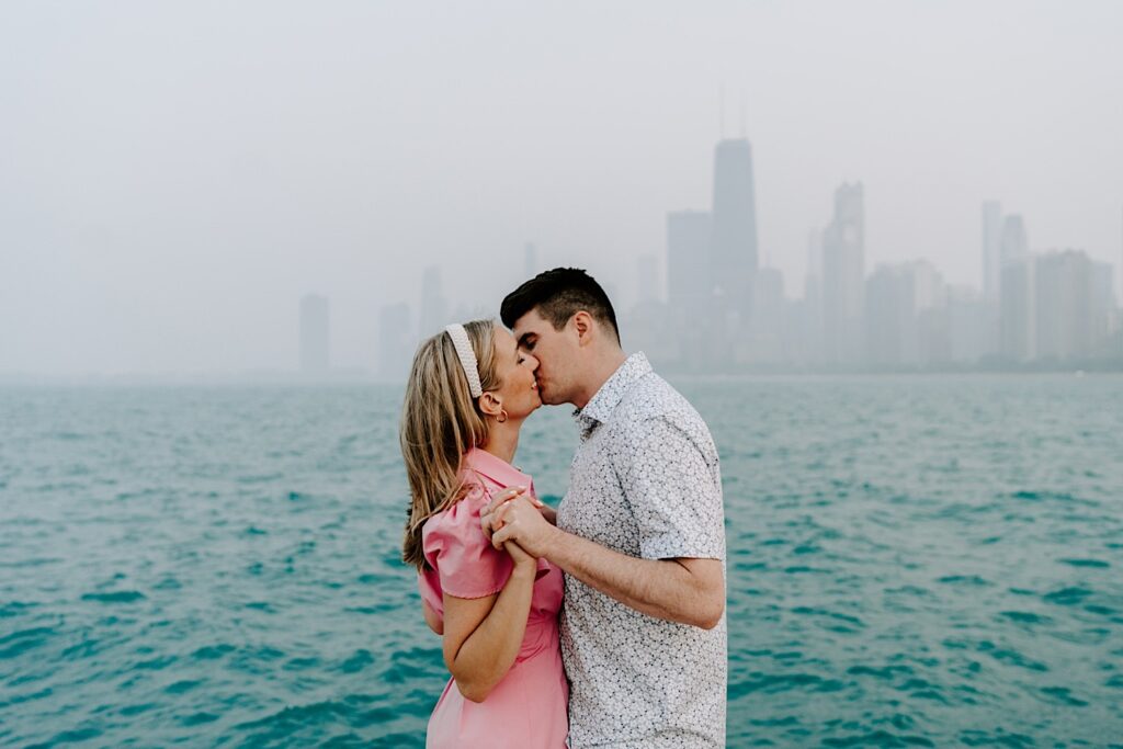 A man and woman kiss one another while standing in front of Lake Michigan and a hazy Chicago skyline during their engagement session