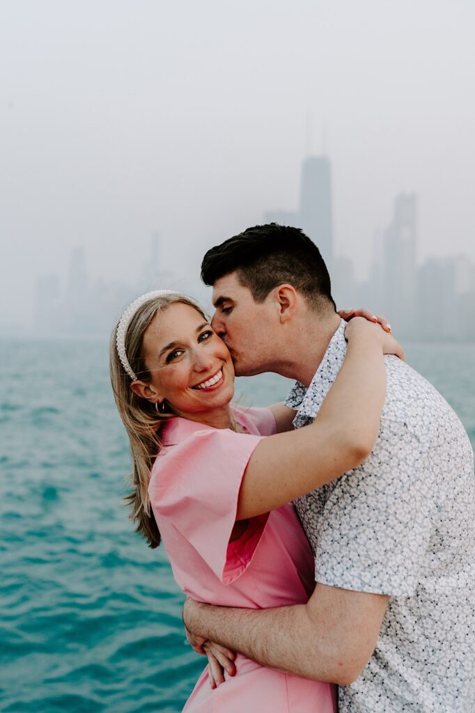 A woman smiles at the camera while hugging a man as he kisses her on the cheek with Lake Michigan and the Chicago skyline behind them