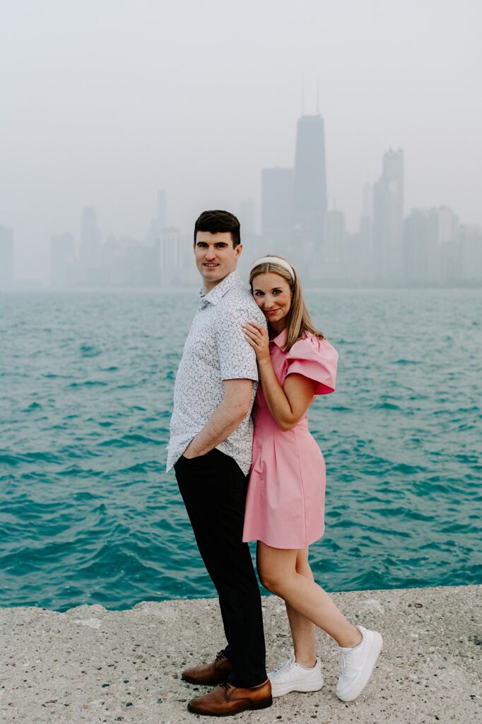 A man and woman look at the camera while the woman stands behind the man and puts her hand on his shoulder while they stand in front of Lake Michigan and the Chicago skyline