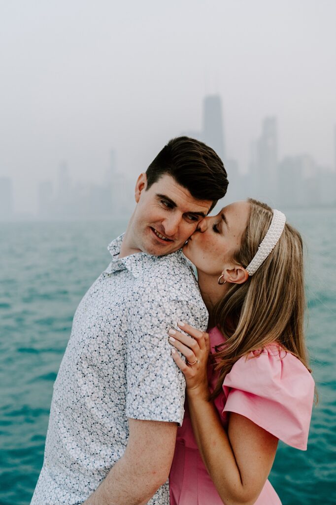 A man looks over his shoulder as a woman behind him kisses him on the cheek in front of Lake Michigan and the Chicago skyline