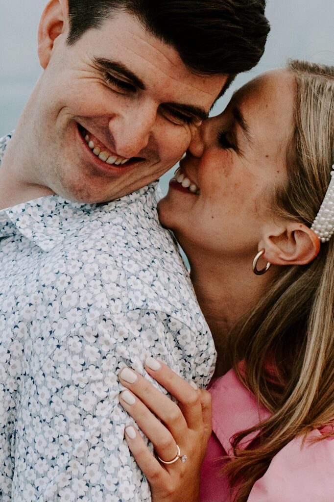 A man smiles over his shoulder as a woman behind him smiles while touching her nose to his cheek during their engagement session