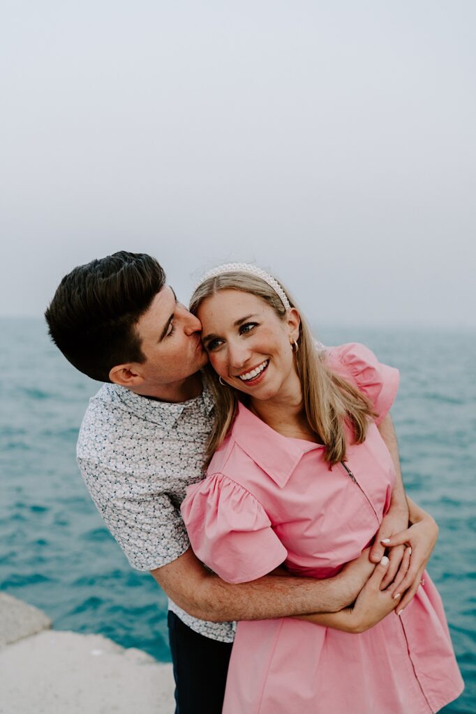 A woman smiles while being hugged from behind by a man who kisses her on her head with Lake Michigan behind them