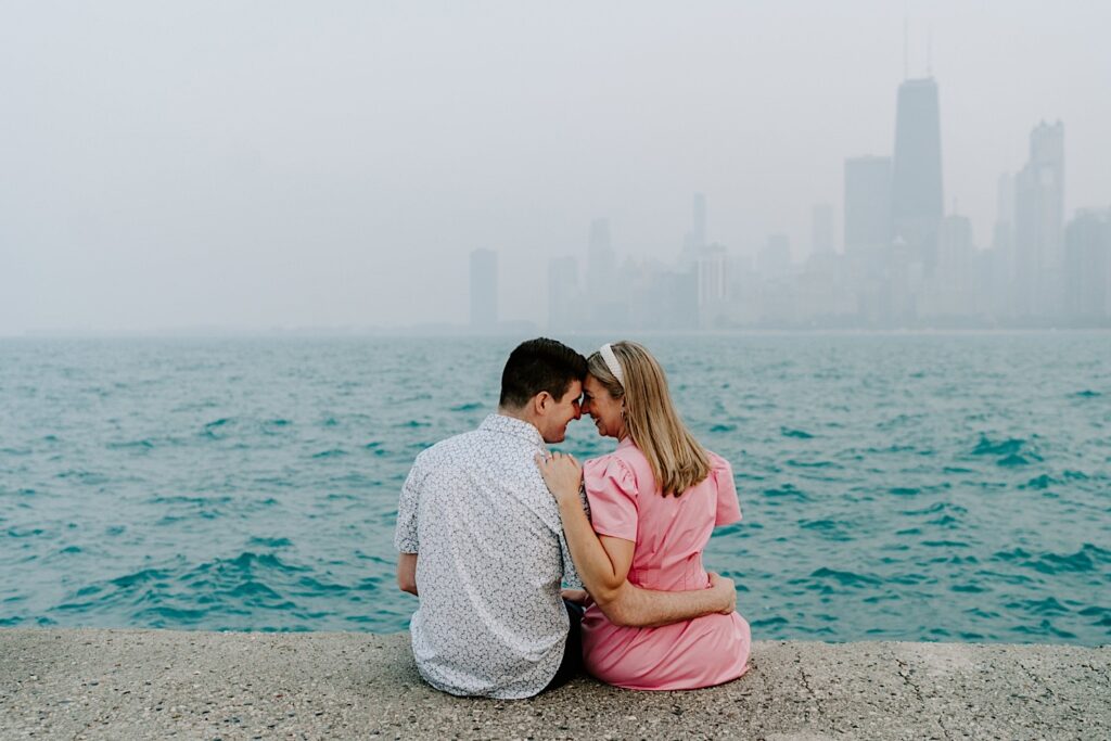 A man and woman sit facing towards Lake Michigan and a hazy Chicago skyline and smile at one another while touching their noses together during their engagement session