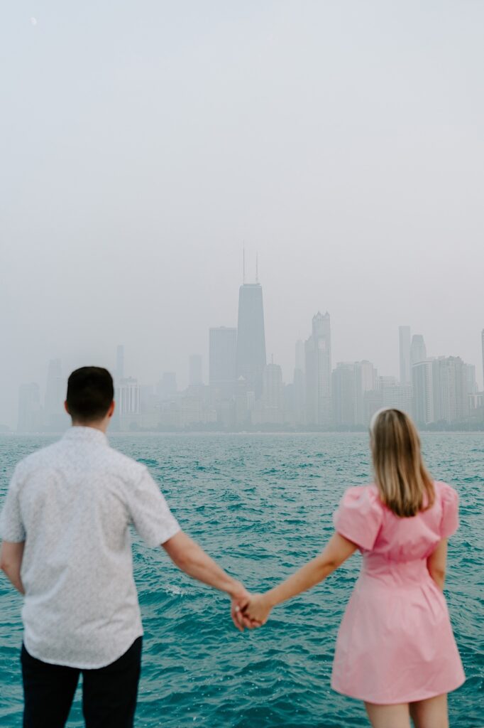 A man and woman hold hands and look out over Lake Michigan towards a hazy Chicago skyline as they face away from the camera