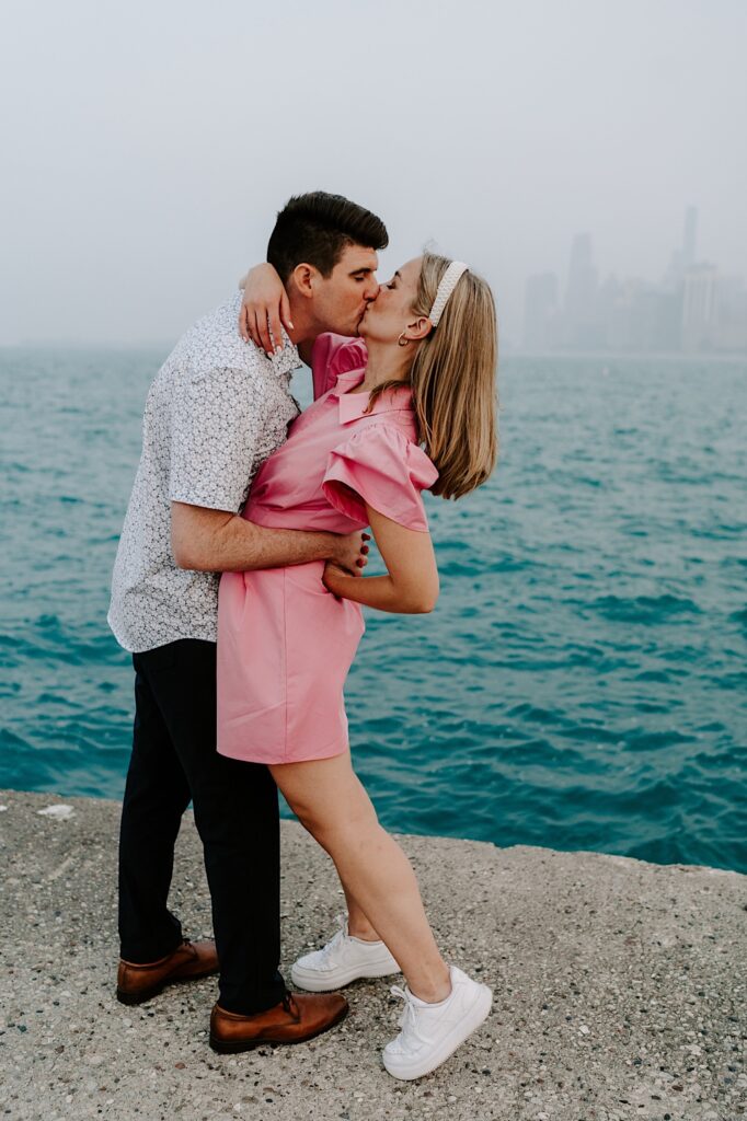 A man and woman kiss one another and embrace while standing in front of Lake Michigan and a hazy Chicago skyline