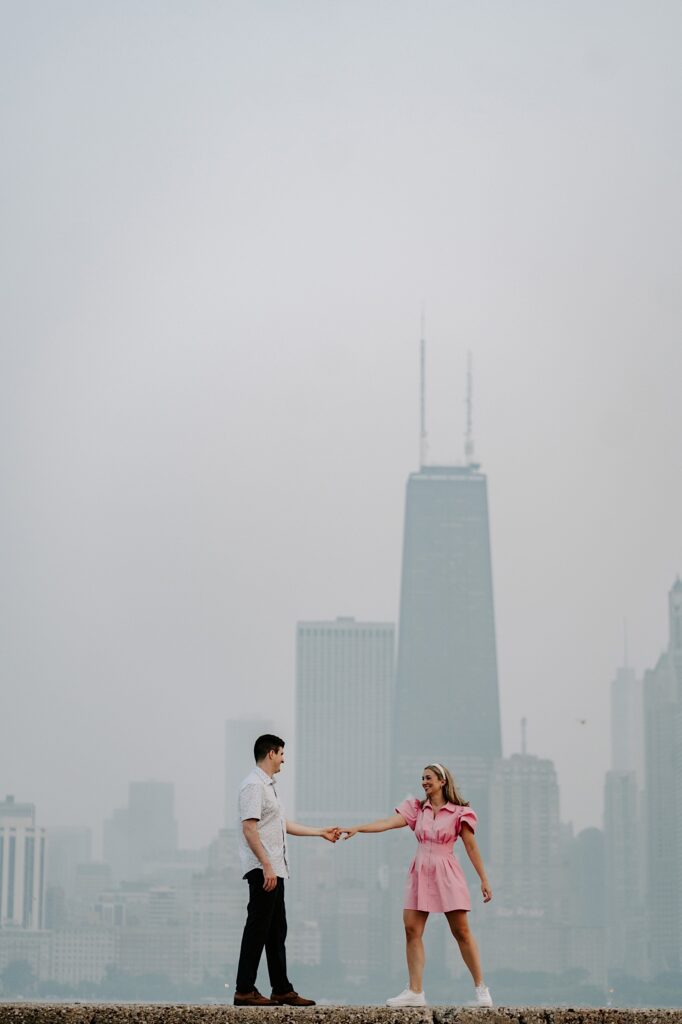 A man and woman hold hands and face one another with a hazy Chicago skyline behind them