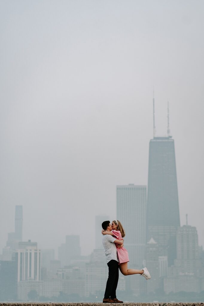 A man picks up a woman and they kiss one another with a hazy Chicago skyline behind them