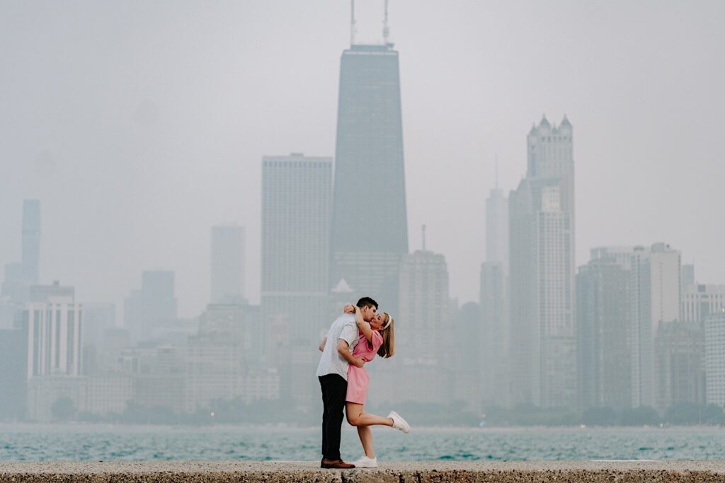 A woman lifts her leg while a man kisses her on the cheek during their engagement session with Lake Michigan and a hazy Chicago skyline behind them