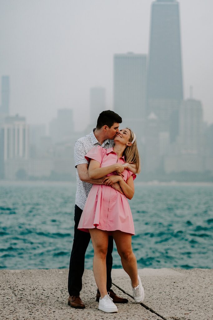 A woman smiles while looking up as a man hugs her from behind and kisses her on the cheek with Lake Michigan and the Chicago skyline behind them
