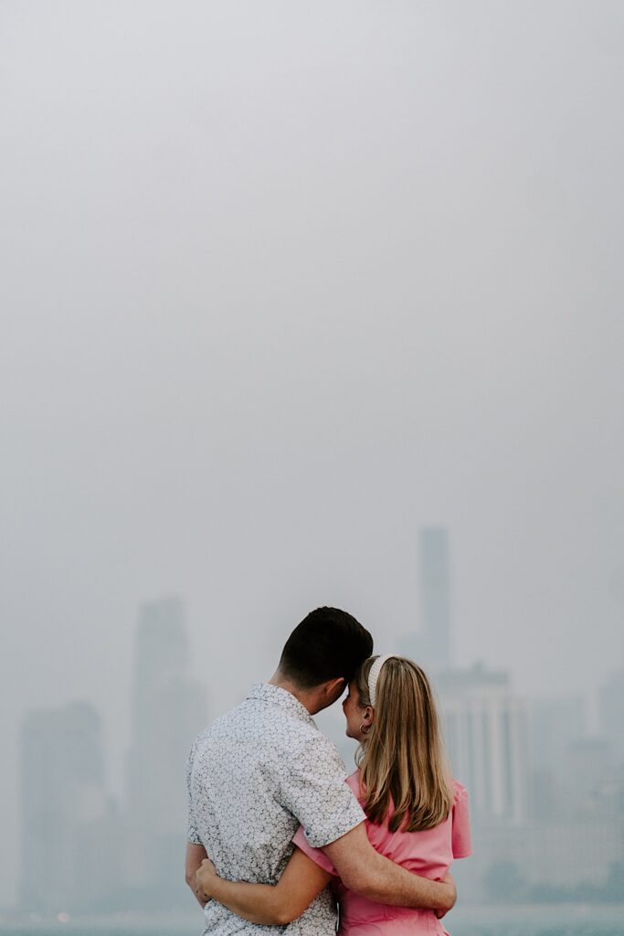 A man and woman face away from the camera and wrap their arms around one another with a hazy Chicago skyline behind them