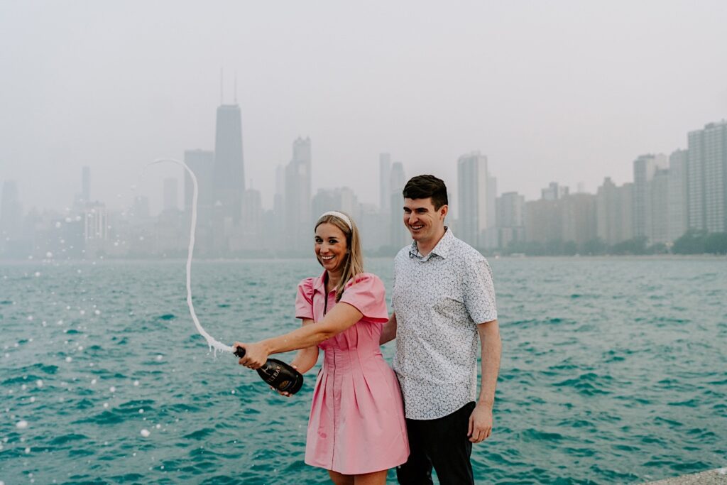 A man and woman smile while standing in front of Lake Michigan and a hazy Chicago skyline during their engagement session, the woman is spraying a bottle of champagne