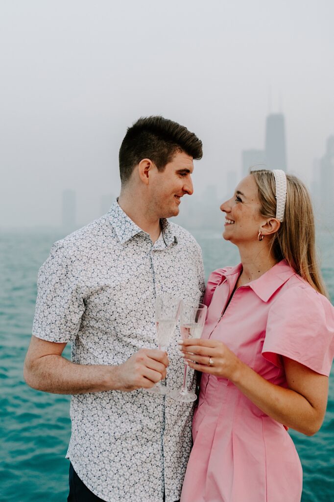 A man and woman smile at one another while each holding a glass of champagne in front of Lake Michigan and a hazy Chicago skyline