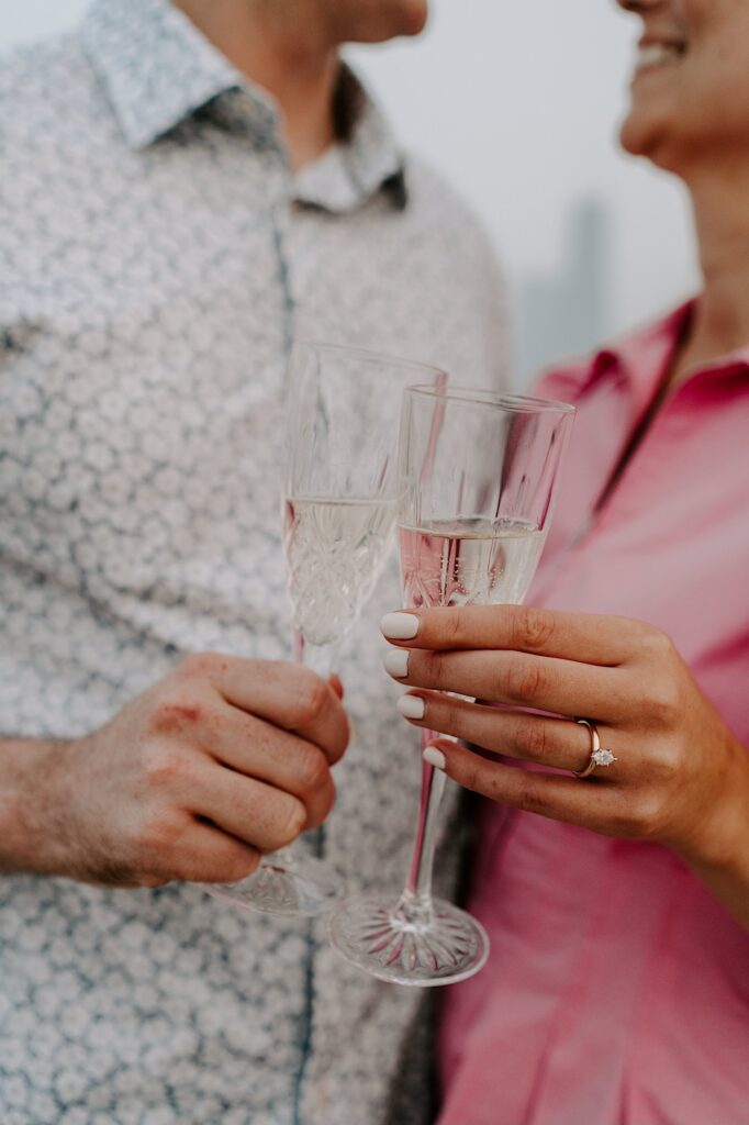Close up photo of a man and woman holding glasses of champagne and smiling at one another, the woman's hand has an engagement ring on it