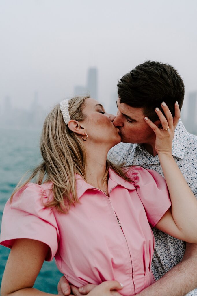 A woman looking over her shoulder kisses a man as he hugs her from behind with Lake Michigan and the Chicago skyline behind them