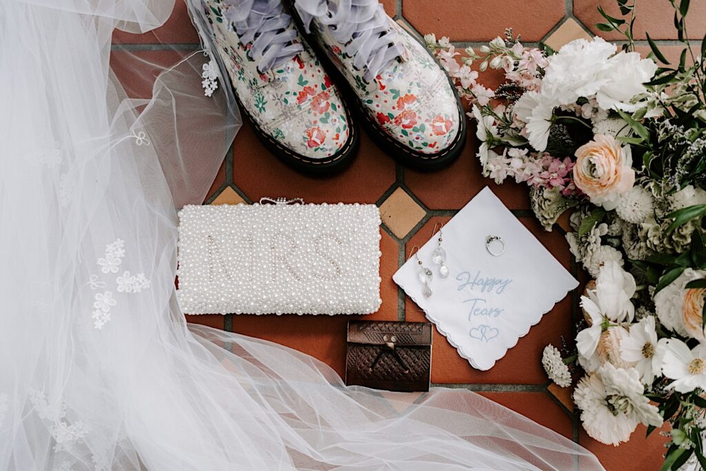 A wedding flatlay on a tile floor with wedding shoes, flowers, a veil, a pearl handbag, earrings, and a wedding ring