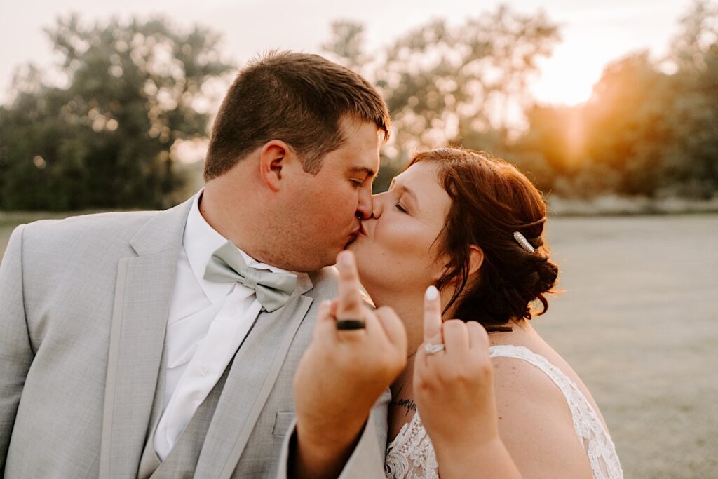 A bride and groom kiss one another while showing off their ring fingers to the camera as the sun sets behind them, photo taken by a Chicago wedding photographer