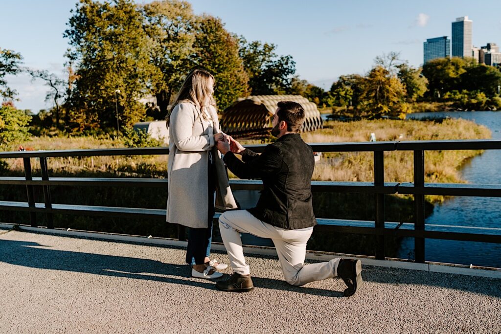A woman holds the hands of a man as he drops to one knee in front of her to propose while they stand on a bridge in Lincoln Park of Chicago