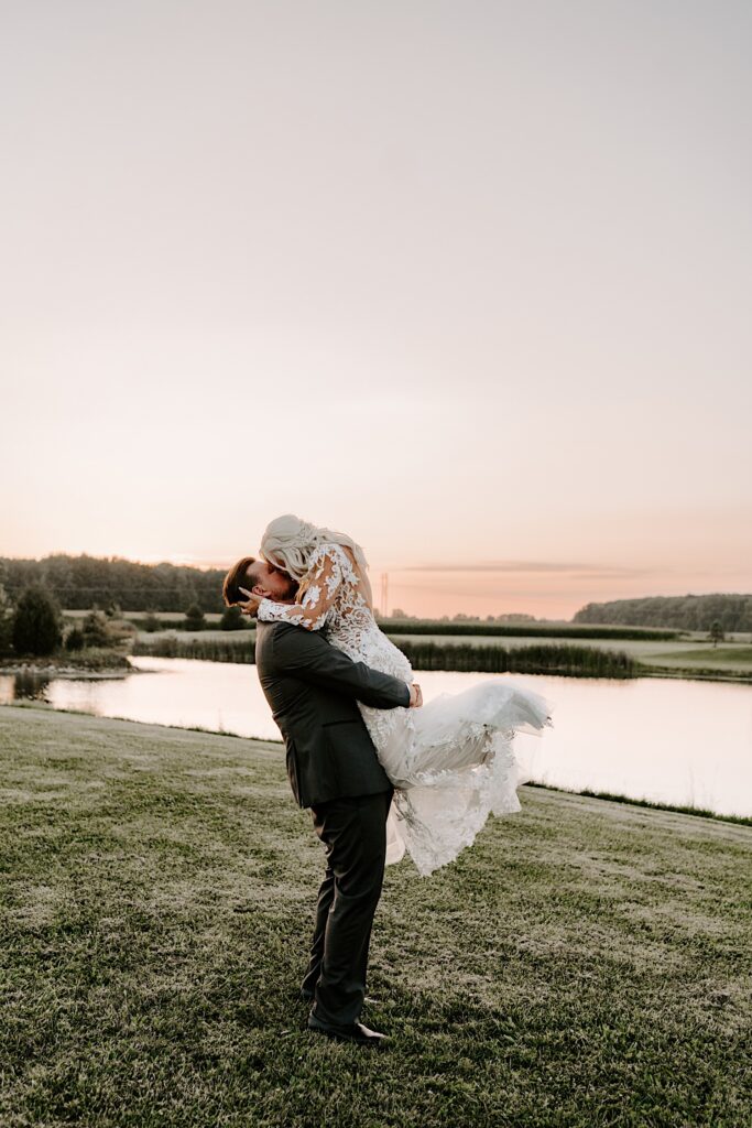 A bride is lifted in the air and kissed by the groom while they stand in a field in front of a pond as the sun sets behind them