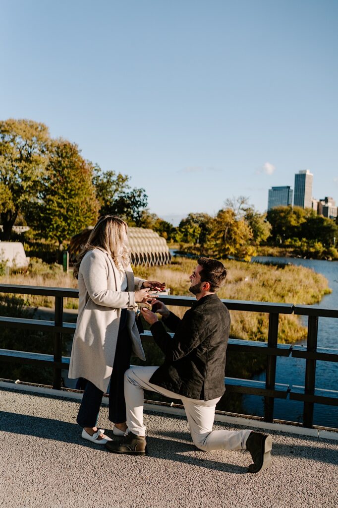 A man down on one knee proposes to a woman while they stand on a bridge in a park in Chicago