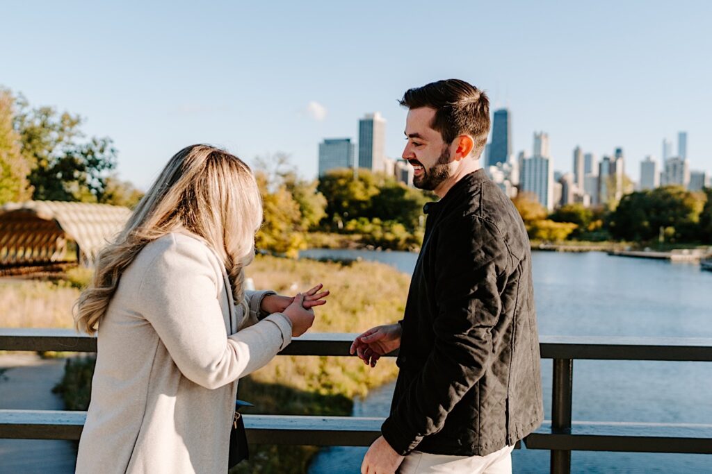 A man smiles as the woman in front of him looks at the ring on her finger after a proposal on a bridge in Lincoln Park of Chicago with the city's skyline in the background