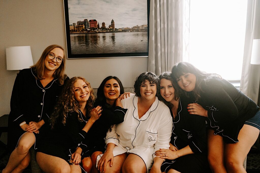 A bride sits on a couch with her 5 bridesmaids as they smile at the camera while in a hotel before they get ready for the bride's wedding day
