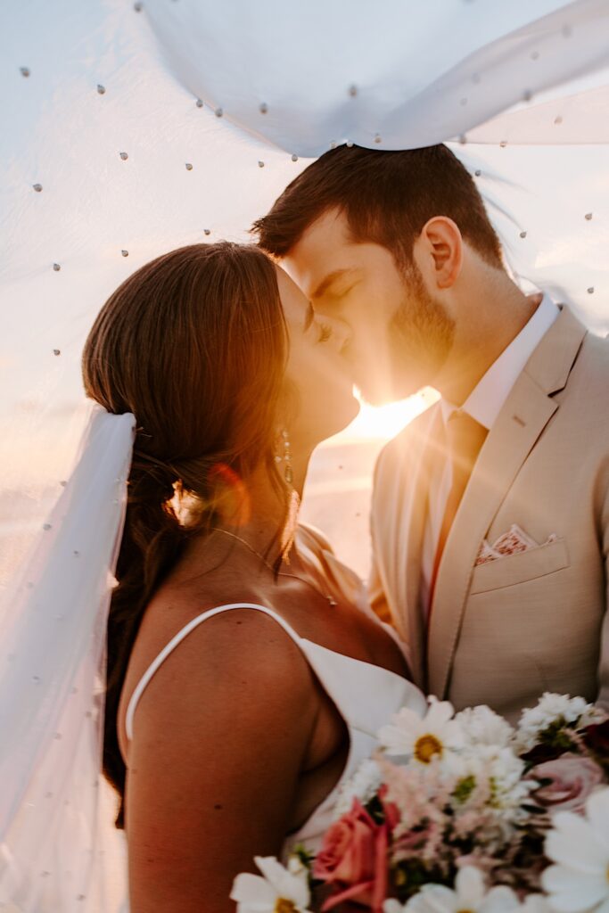 A bride and groom kiss one another while underneath the bride's veil as the sun sets behind them