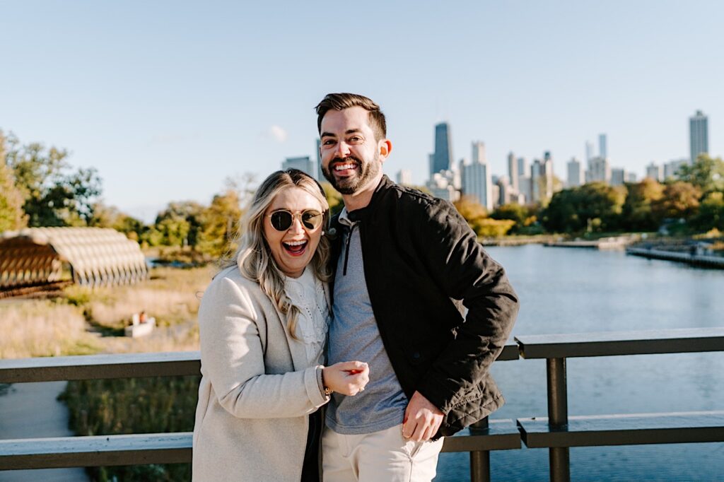 A man and woman embrace and smile at the camera after a proposal while standing on a bridge in Lincoln Park, behind them is a lake and the Chicago skyline