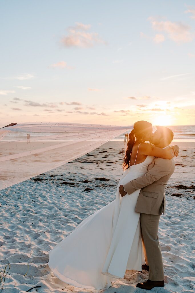 A bride and groom stand on a beach together and kiss as the groom lifts the bride in the air, behind them the sun sets on the ocean
