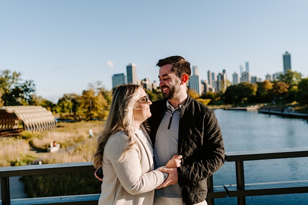 A man and woman embrace and smile at one another while standing on a bridge in Lincoln Park after their proposal, behind them is a lake and the Chicago skyline
