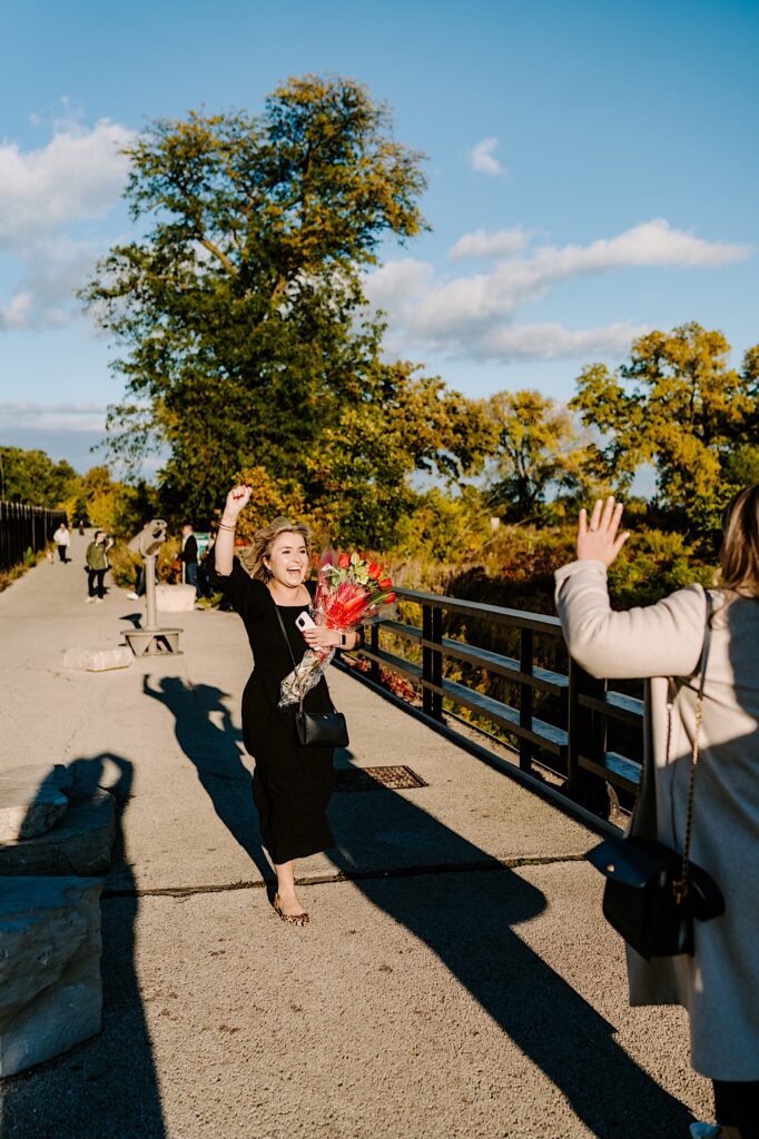 A woman carrying flowers cheers as she approaches another woman on a bridge after a proposal