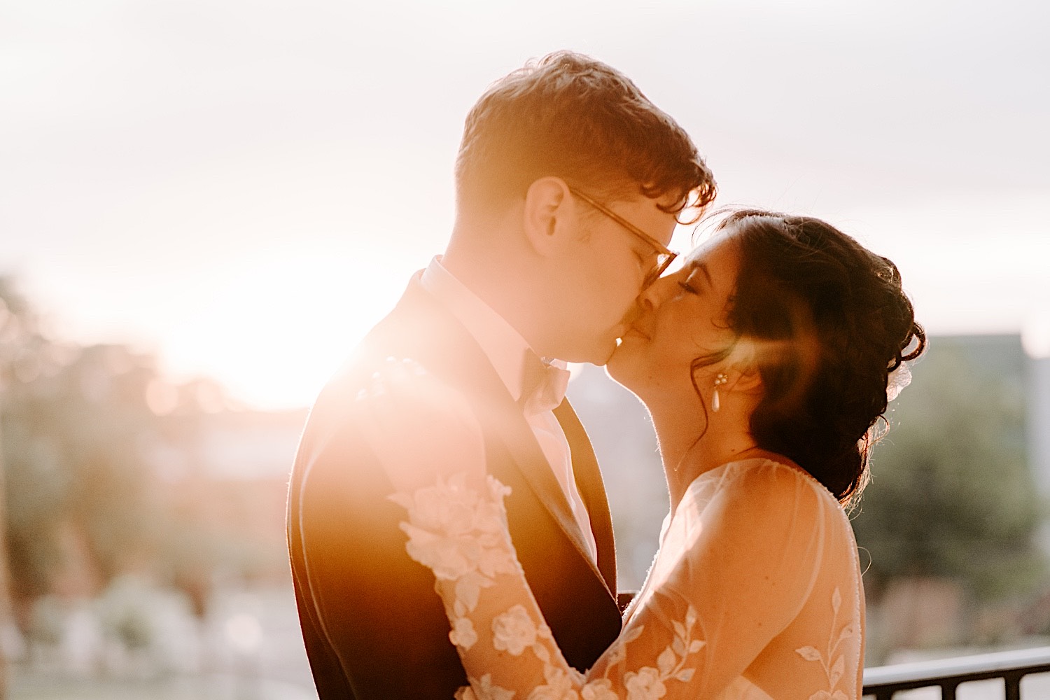 A bride and groom stand on a balcony and kiss one another as the sun sets directly behind them, photo taken by a Chicago wedding photographer