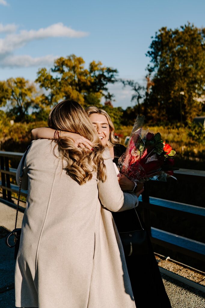 Two women hug while standing on a bridge, one holds flowers that she brought for the other woman who just was proposed to