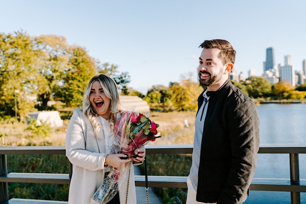A woman smiles while holding flowers and standing next to a man who is also smiling after their proposal on a bridge in Lincoln Park, behind them is a lake and the Chicago skyline