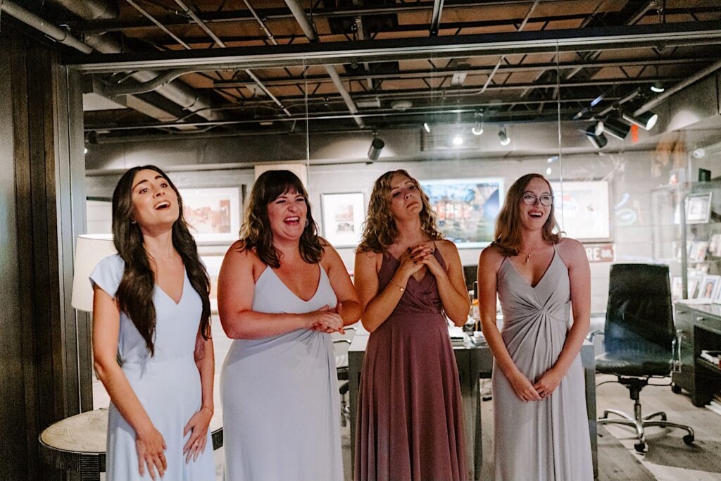 Bridesmaids stand together and react with joy seeing the bride who is off camera in her wedding dress for the first time