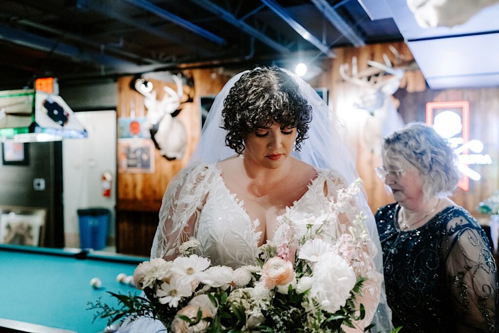 A bride stands with her flower bouquet and looks down at it while her mother stands behind her