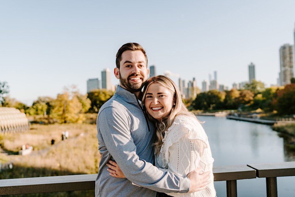 A man and woman hug one another and smile at the camera after their proposal while standing on a bridge in Lincoln Park with a lake and the Chicago skyline behind them