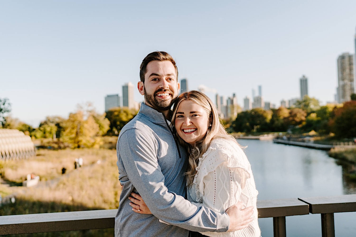 A man and woman hug one another and smile at the camera after their proposal while standing on a bridge in Lincoln Park with a lake and the Chicago skyline behind them