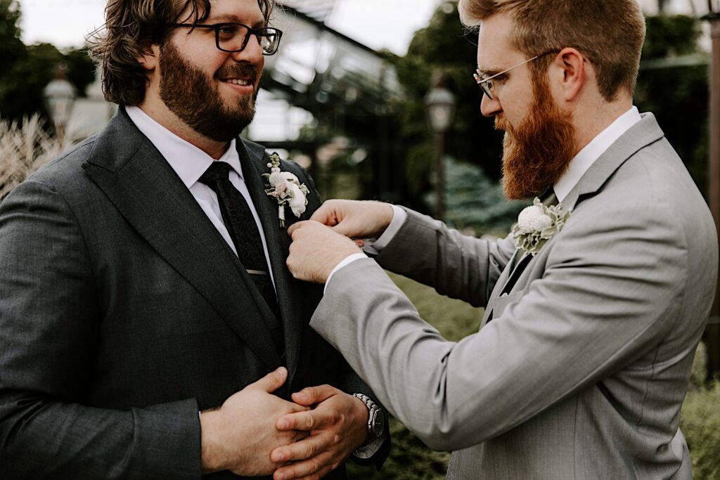 A groom smiles while standing outside as a groomsmen helps pin his boutonniere to his suit