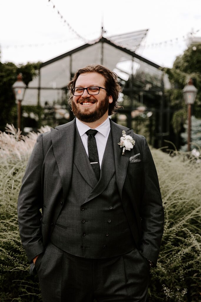 A groom smiles while in his wedding suit standing outside with his hands in his pockets