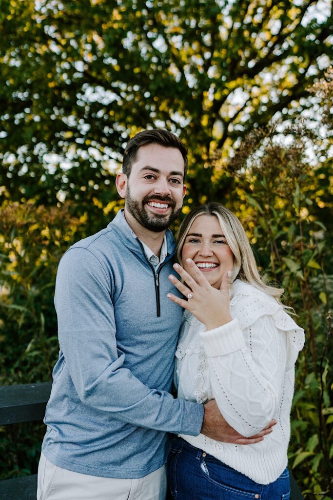 A couple smile at the camera as the woman shows off her engagement ring while they stand in front of some of the plants that make up Lincoln Park