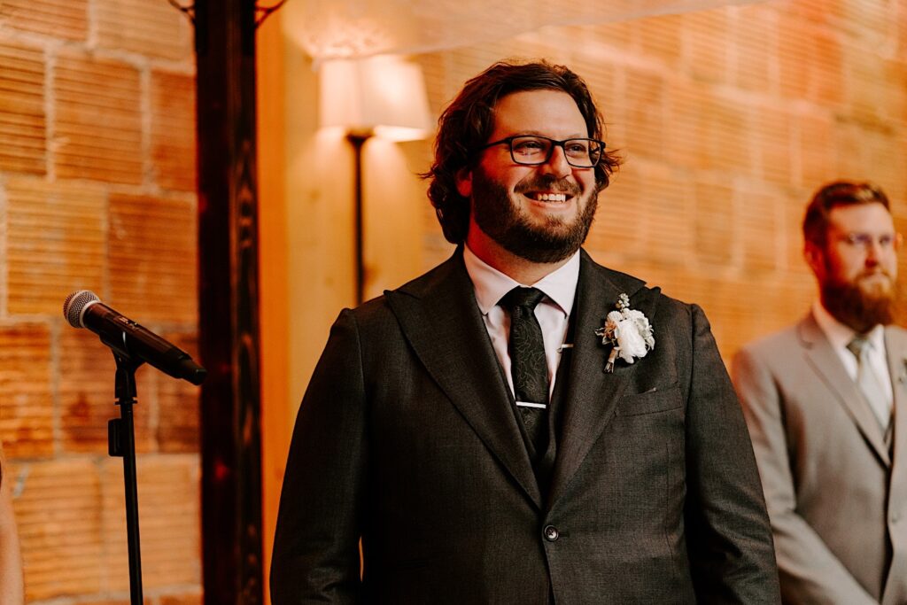 A groom smiles while standing in front of a brick wall during his wedding ceremony at The Atrium in Milwaukee
