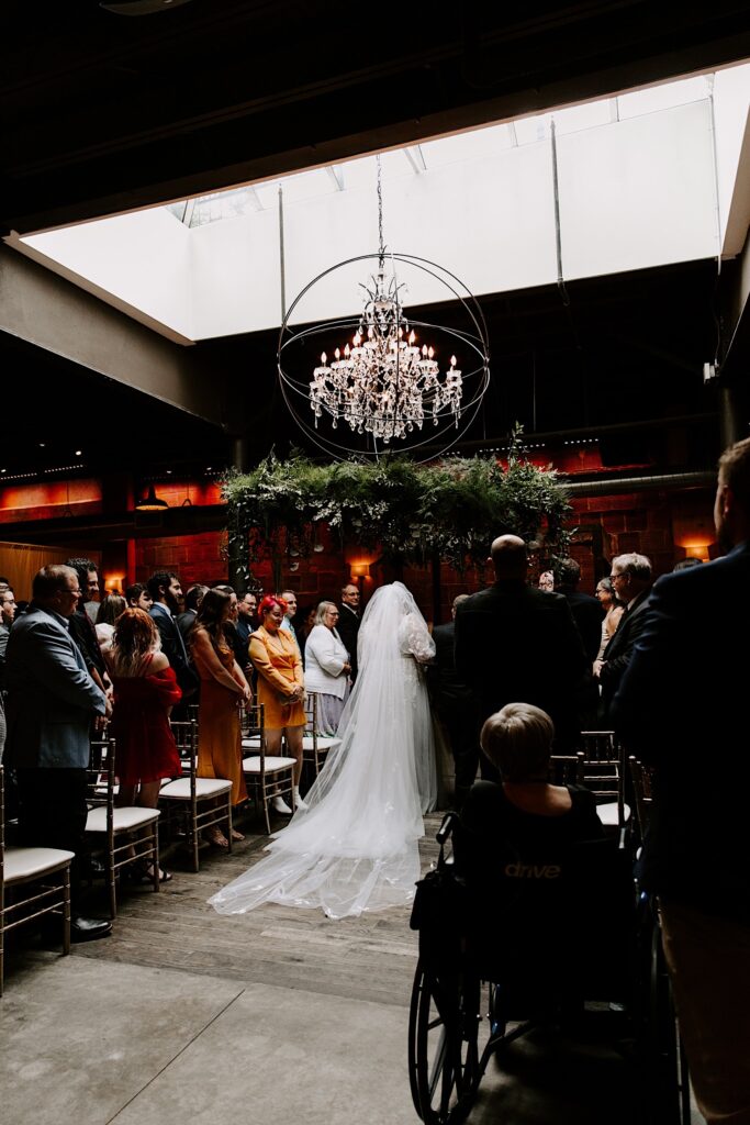 A bride walks down the aisle with her father for her wedding ceremony underneath a large glass chandelier hanging from a skylight
