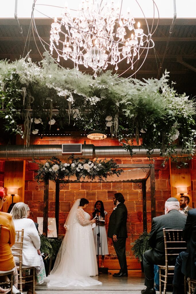 A bride and groom read their vows to one another during their indoor wedding ceremony underneath a skylight and a large chandelier