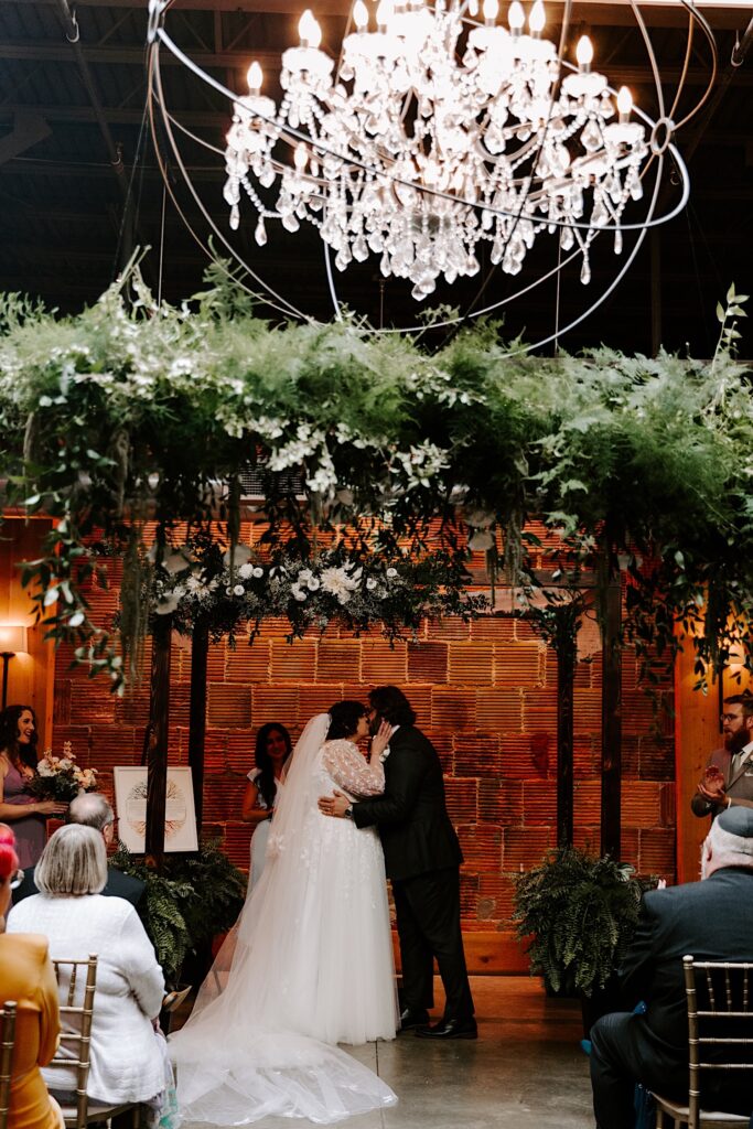 A bride and groom kiss one another during their wedding ceremony while underneath a large chandelier