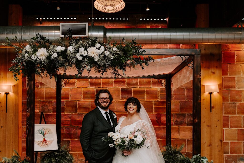 A bride and groom stand side by side and smile at the camera in their wedding ceremony space which is inside The Atrium in Milwaukee in front of the venue's brick wall