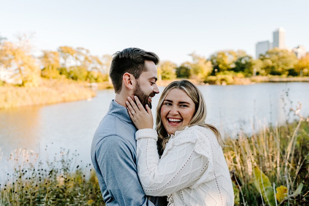 A woman smiles at the camera while holding the face of a man who smiles back at her as the two stand in front of a lake in Lincoln Park after their proposal